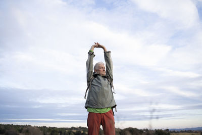 Senior male hiker stretching arms while standing against cloudy sky at countryside