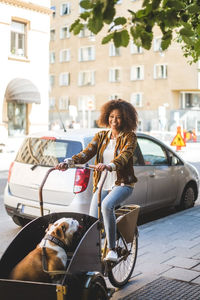 Full length of happy woman riding bicycle cart with bulldog on sidewalk by car in city