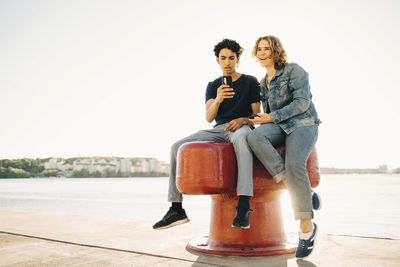 Full length of teenage boy using mobile phone by cheerful friend on bollard against clear sky in city