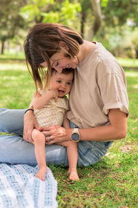 Positive mom sitting on lawn with adorable little daughter while spending pleasant time in park