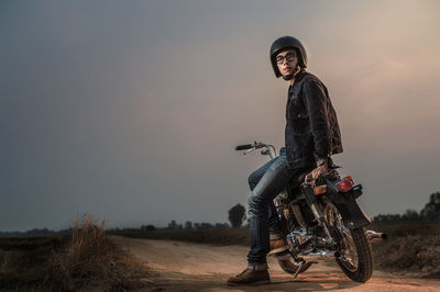 Thoughtful young man sitting on motorcycle at field against sky