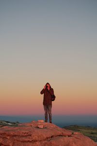 Full length of man photographing through camera while standing on rock during sunset