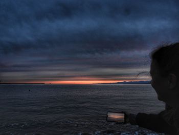 Side view of man photographing sea against sky during sunset