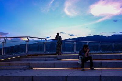 Man photographing while woman standing by railing against sky