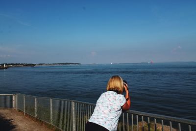 Woman photographing while standing by railing against sea