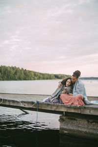 Daughter showing smart phone to mother while sitting on pier over lake during sunset