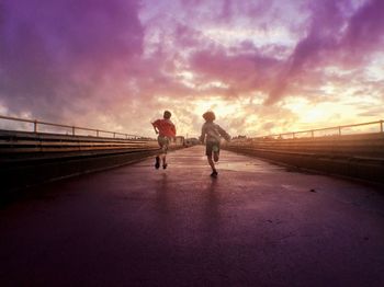 Rear view of boys running on walkway against cloudy sky at sunset