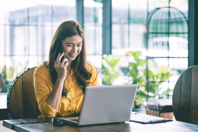 Smiling businesswoman using laptop while talking on mobile phone in office