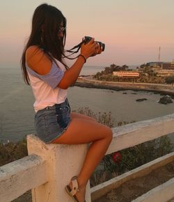 Young woman photographing sea against sky during sunset