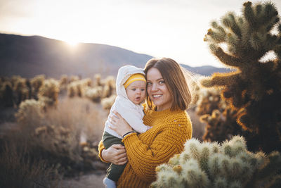 A woman with a baby is standing near a cactus in the desert