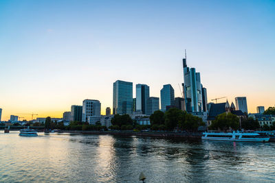 Modern buildings by river against sky during sunset