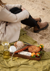 Unrecognizable female in elegant coat sitting on blanket near bottle and glass of white wine and tray with pie with tangerines and bread with cheese and sausages while resting on beach on weekend day