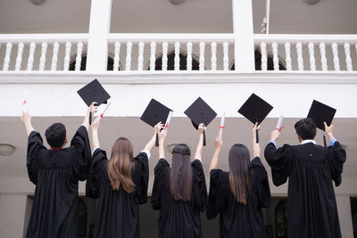 Rear view of students in university gowns holding mortarboards while standing against building