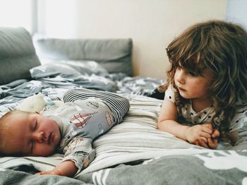 Cute girl looking at brother sleeping on bed at home