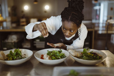 Focused female chef sprinkling salt on salad while bending in commercial kitchen