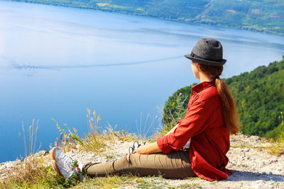 Young woman in hat looks on the sea solo female sitting on the shore of an mountain lake dreamily. 