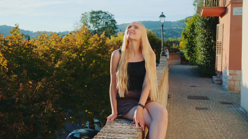 Young woman sitting on sidewalk against trees