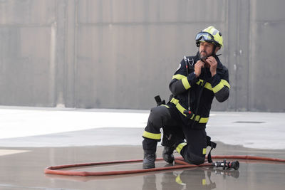 Contemplative unshaven firefighter in uniform with stripes and protective helmet reflecting in cement surface near hose at work while looking away