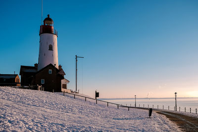 Lighthouse by snow covered building against sky during sunset