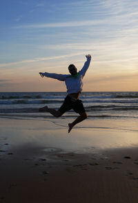 Silhouette young man with arms outstretched jumping at beach against sky during sunset