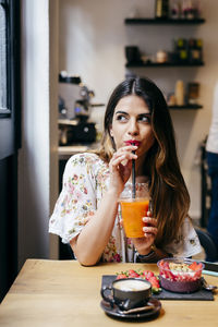 Beautiful cheerful modern female with stylish earrings enjoying fruit juice while having breakfast sitting at wooden table in cafe