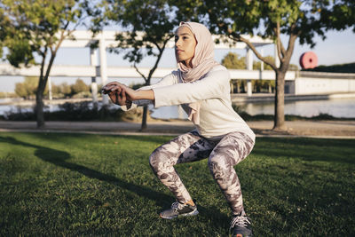 Full body of determined muslim female in hijab doing squat extending arms forward for balance during outdoor training