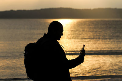 Silhouette man using phone by sea during sunset