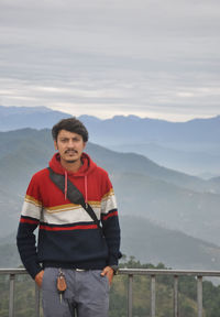 A guy looking at camera with hands in pocket while standing against the background of mountains