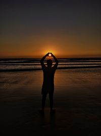 Silhouette man making heart shape with hands at beach against sky during sunset