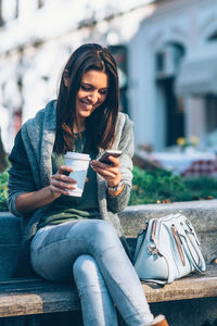 Smiling young woman using mobile phone while holding disposable cup in city