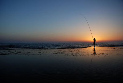 Silhouette man fishing on beach against sky during sunset