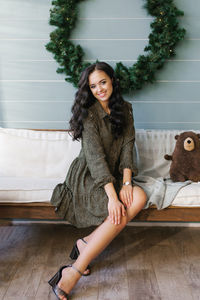 Young woman in a dress is sitting on a white sofa against the background of a large christmas wreath