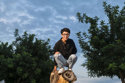 Young transgender man with glasses posing on a log outdoors.