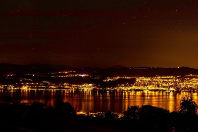 Illuminated cityscape by river against sky at night