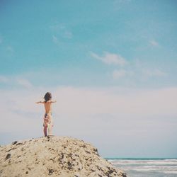 Shirtless boy with arms outstretched standing on rock at beach against sky