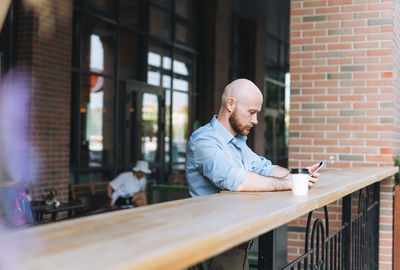 Adult attractive man forty years with beard in blue shirt businessman using mobile phone at cafe