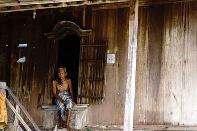 Full length of shirtless man sitting by wooden house