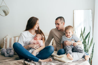 Mom and dad are sitting on the bed with their newborn daughter and toddler son.