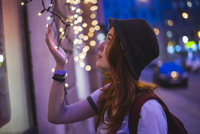 Side view of stylish hipster millennial woman in hat and with backpack touching glowing garland while standing near building in evening city