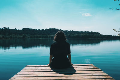 Rear view of woman sitting on pier over lake against sky