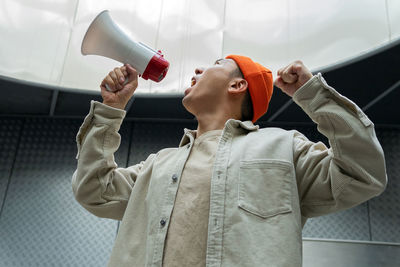 Side view of overwhelmed male protesting in casual clothes screaming in loudspeaker while standing against gray wall in dark room