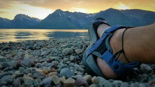 Low section of man in wearing sandal at beach against sky during sunset