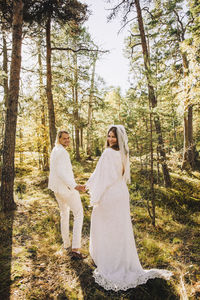 Full length of smiling newlywed bride and groom holding hands looking back in forest
