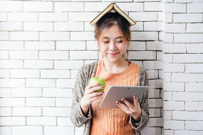 Close-up of young woman with book on head using digital tablet while holding granny smith apple against wall
