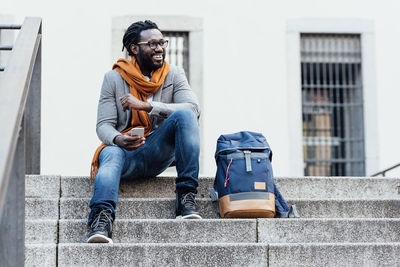 Smiling man using mobile phone while sitting on steps in city