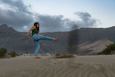 Full length of woman kicking sand against mountains and sky