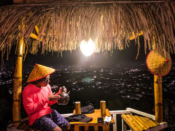 Side view of man sitting at illuminated park against sky at night