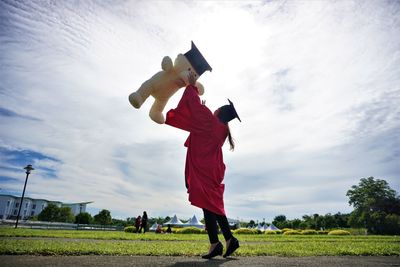 University student throwing teddy bear against sky during graduation