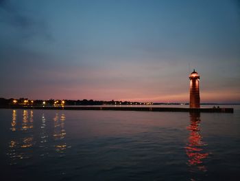 Sea by illuminated building against sky during sunset