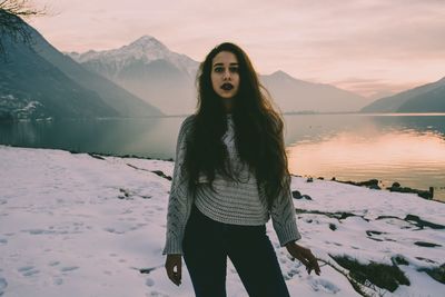 Portrait of woman standing by lake against mountain during winter at sunset